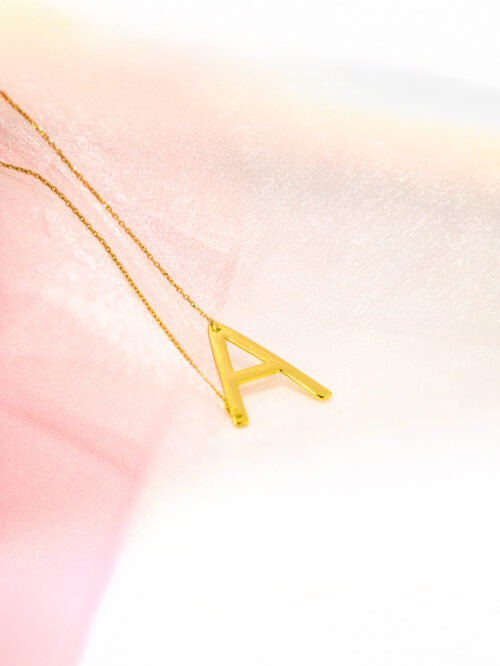 Gold necklace featuring the initial 'A' on a pastel pink and white background.​