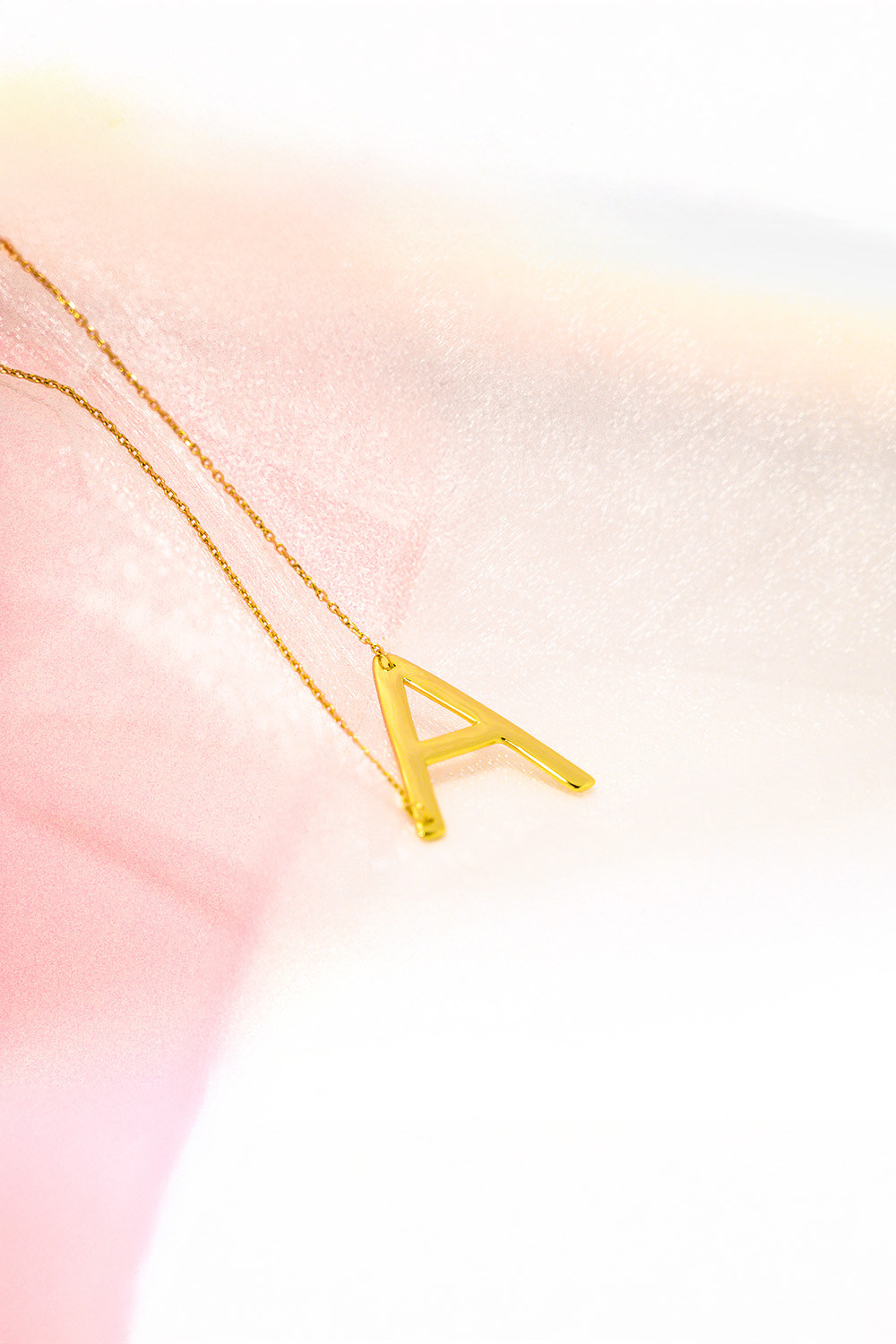 Gold necklace featuring the initial 'A' on a pastel pink and white background.​